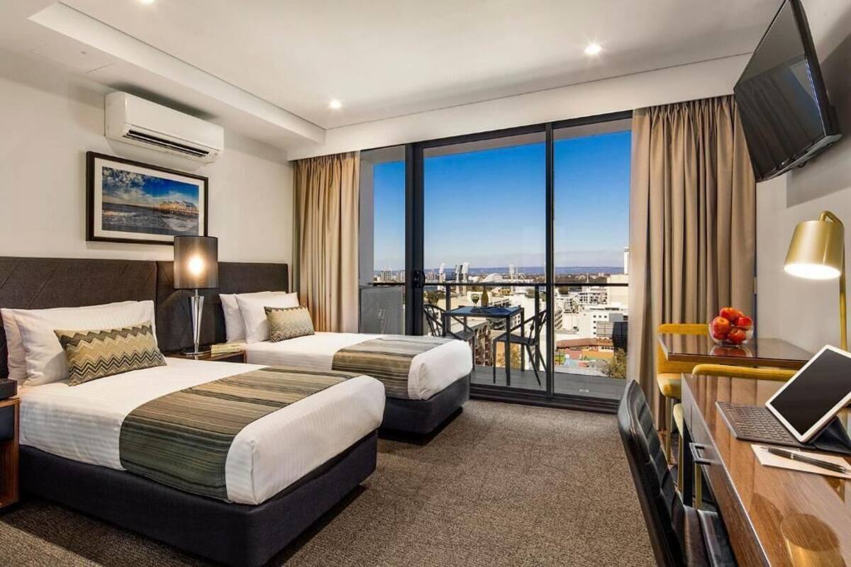A hotel room with two beds, a desk with a laptop, a TV, and a balcony with a city view.