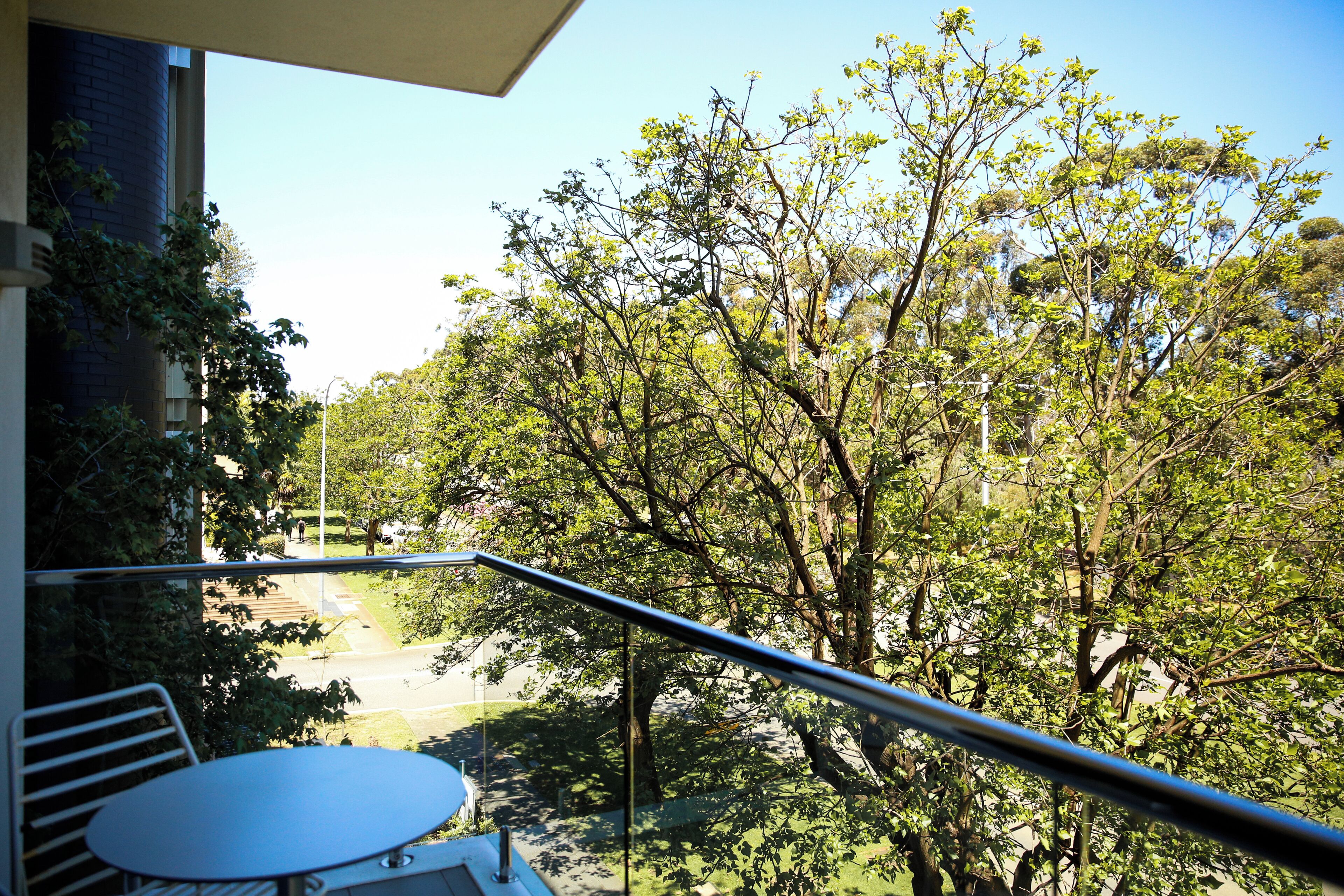 A balcony with a table and chairs, overlooking a street with trees and a clear sky.
