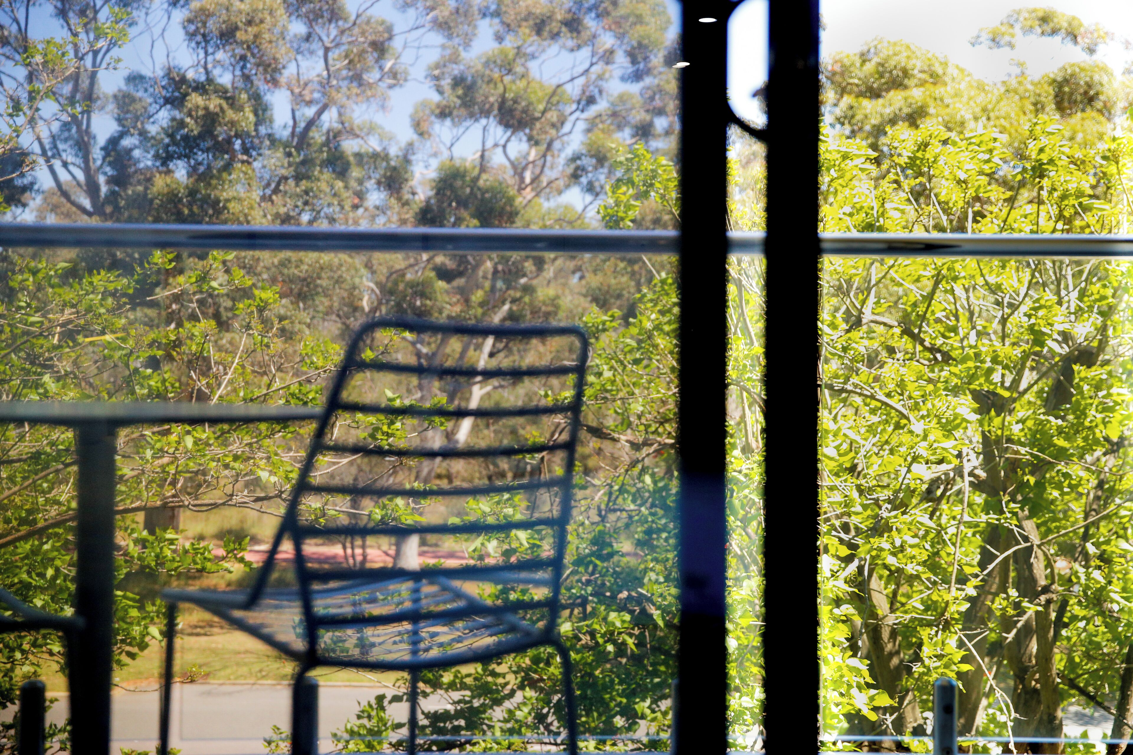 A metal chair with a grid backrest, positioned on a balcony with a view of trees and a clear sky.