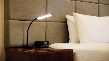Un lit bien fait, avec du linge de lit blanc, une table de chevet sur laquelle se trouvent un réveil noir et une lampe, et une tête de lit au motif capitonné.