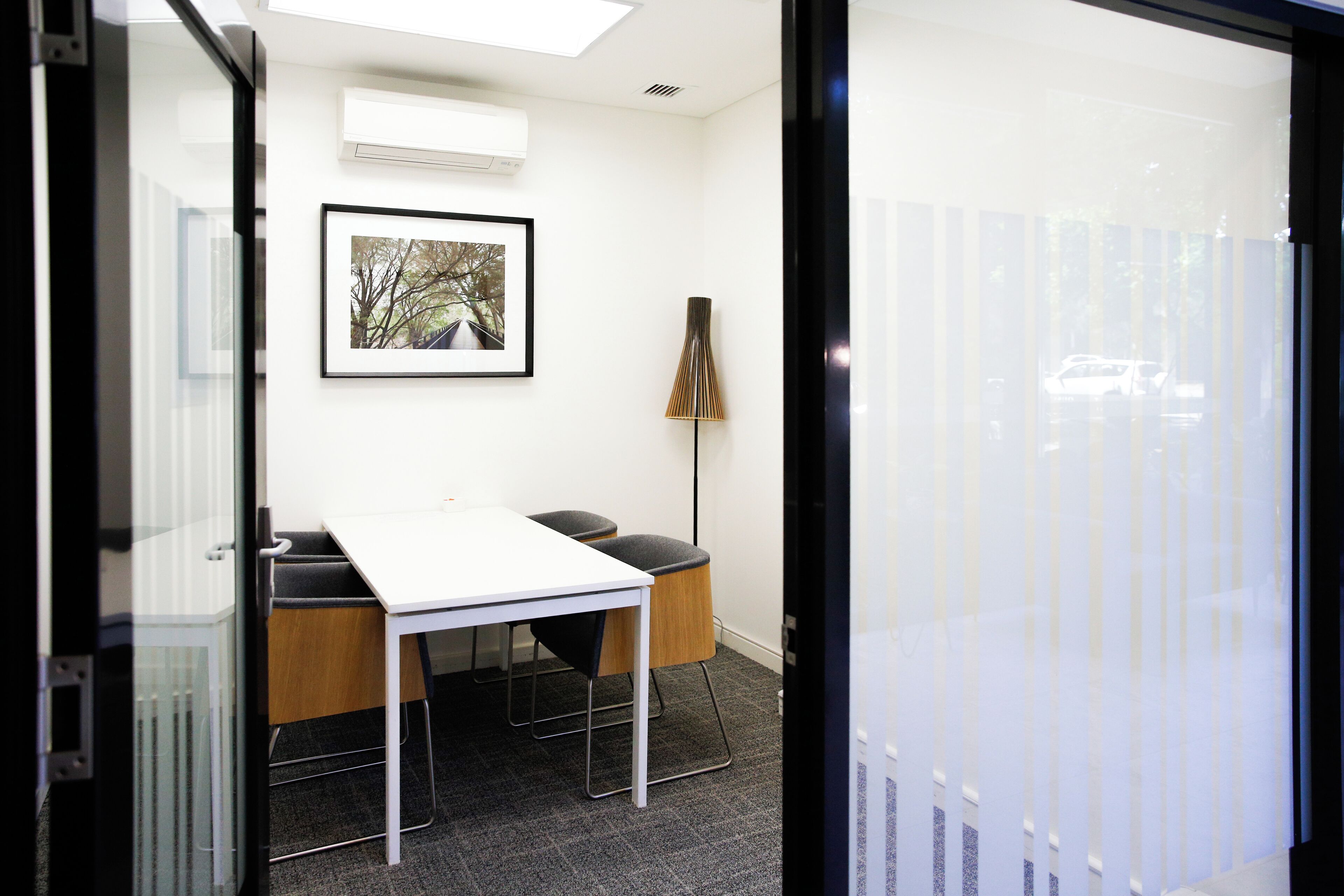 A modern office space with a white table, two black chairs, a framed picture on the wall, and a tall floor lamp.