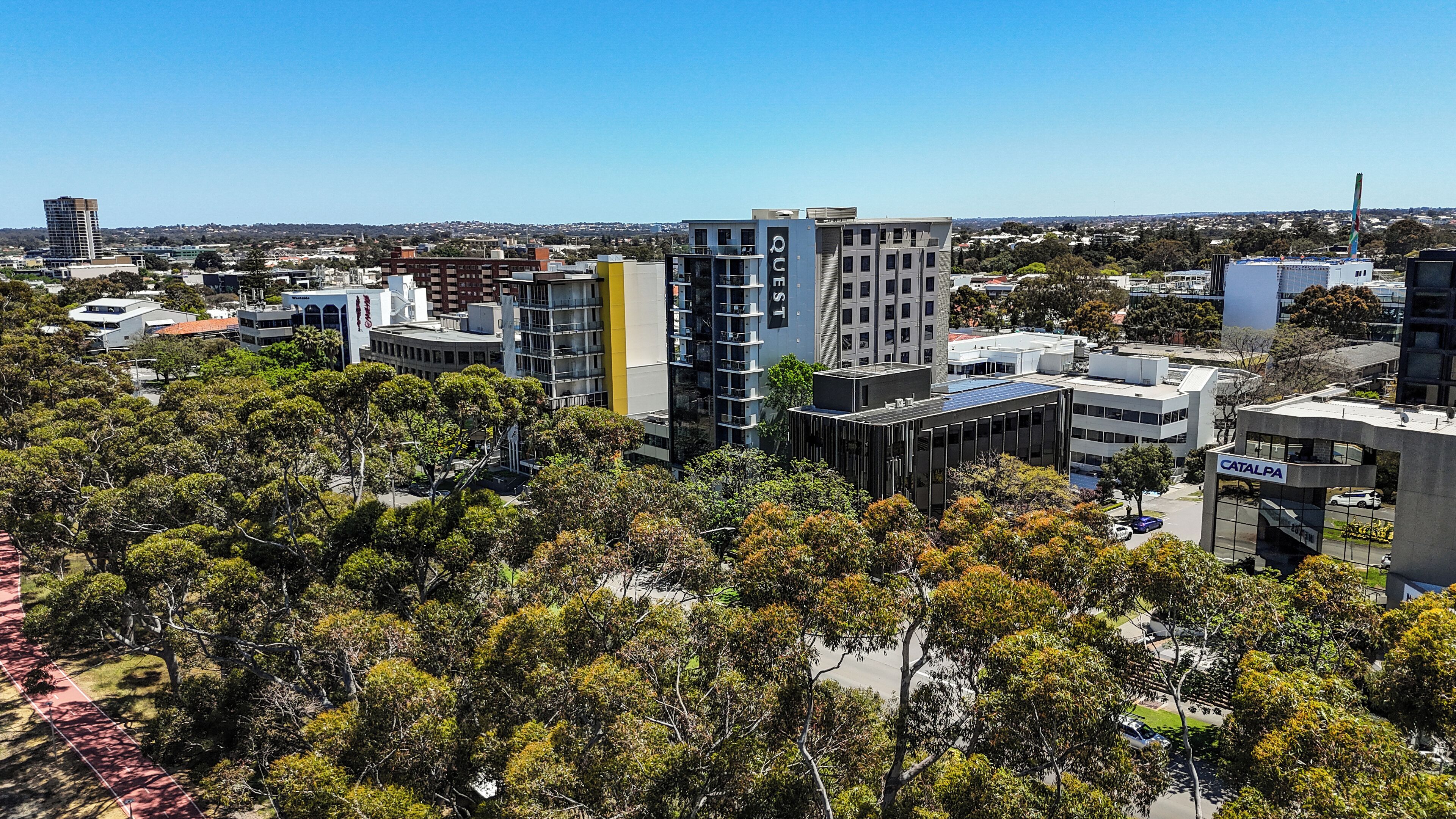 A modern urban area with high-rise buildings, a mix of commercial and residential structures, and a dense tree line in the foreground.