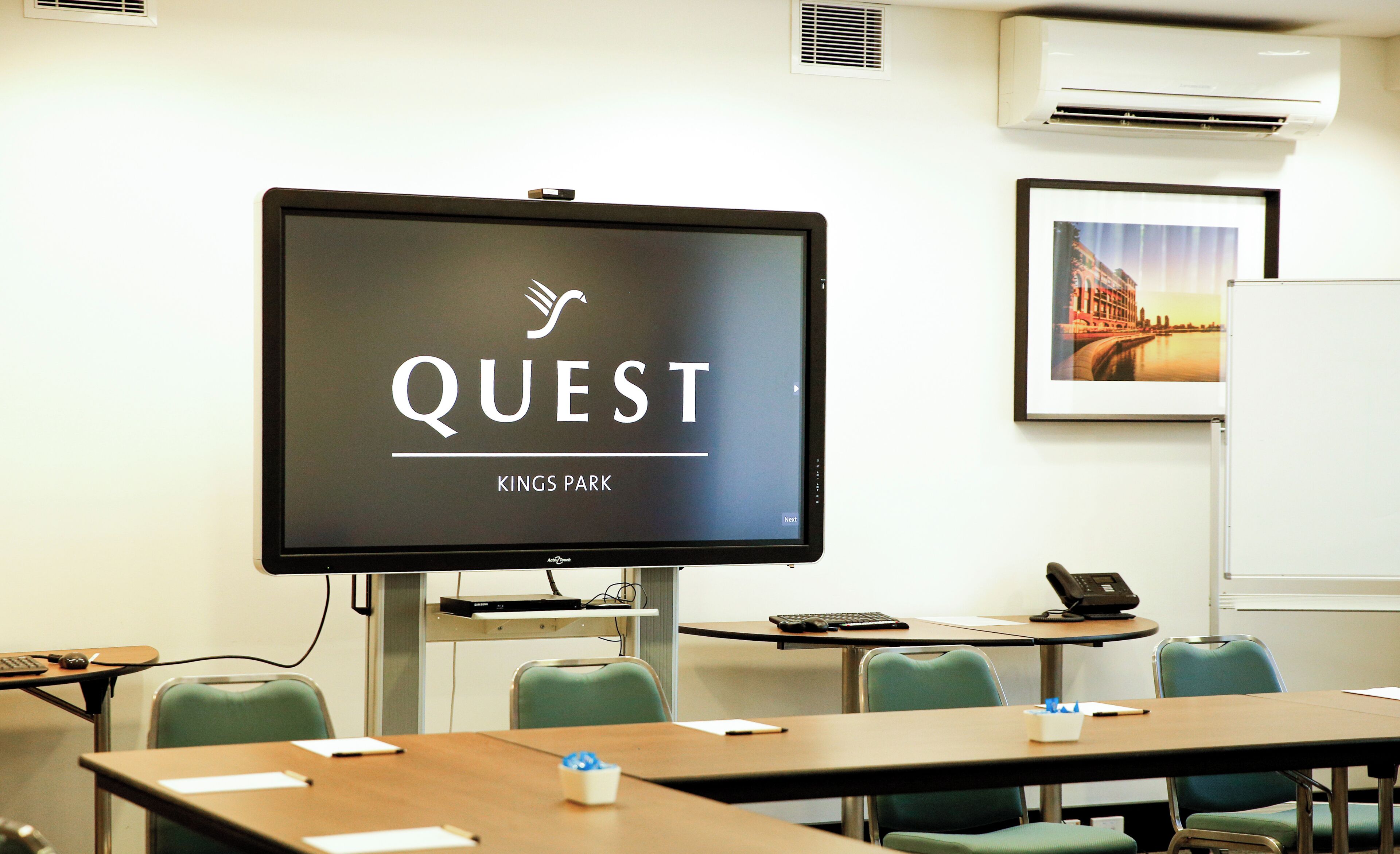 A conference room with a large screen displaying "QUEST KINGS PARK" and a framed picture of a cityscape on the wall.
