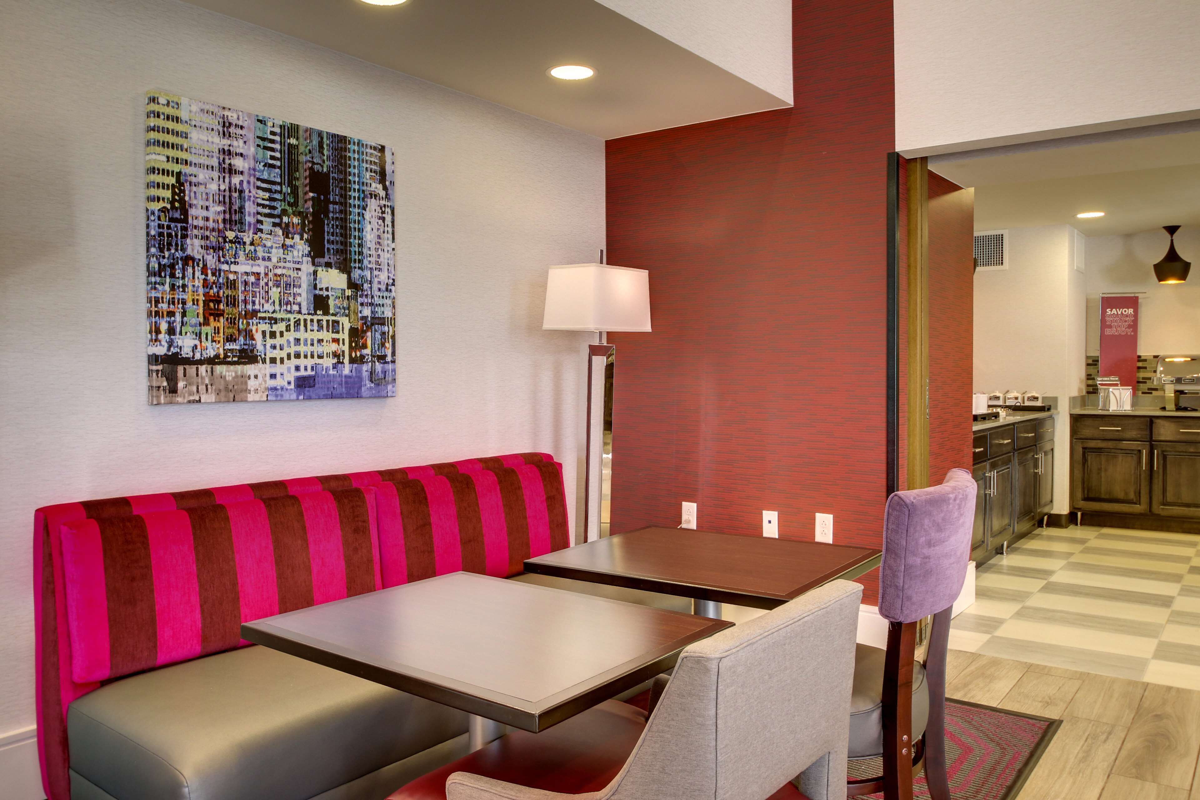 Een moderne hotel lobby met een geruit vloerkleed, een bank met rode en bruine strepen, een tafel met een lamp, en een kleurrijk stadsgezichtschilderij aan de muur.