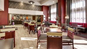 Un hall d’hôtel spacieux, doté de plusieurs espaces salon, d’un bar central et offrant une vue sur la cuisine en arrière-plan.