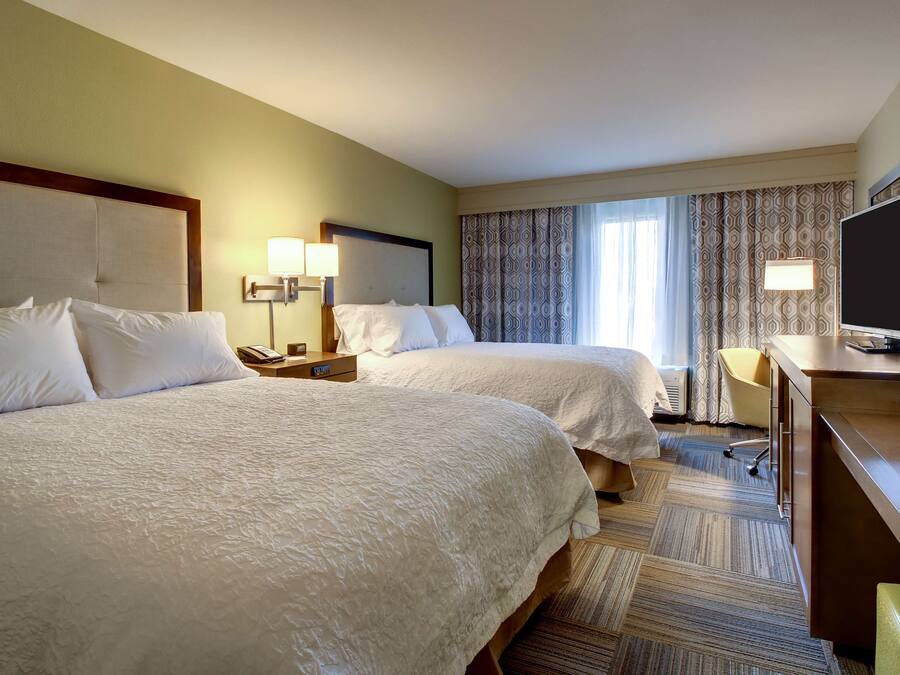 A hotel room with two beds, a desk with a television, a lamp, and a window with curtains.