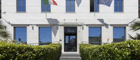A building with a symmetrical facade, large windows, and a central entrance. Flags of Italy and the European Union are displayed.