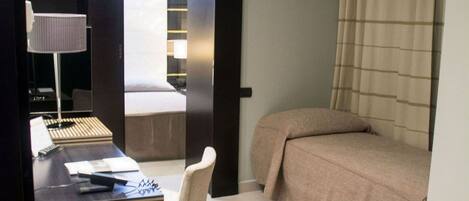 A hotel room with a bed, a desk with a chair, a small stool, and a nightstand with a lamp.