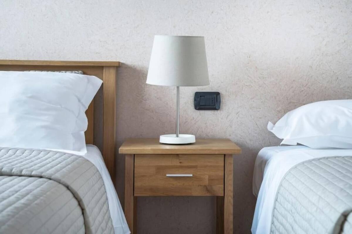 Quarto de hotel com duas camas, uma mesa de cabeceira com abajur e uma tomada na parede.