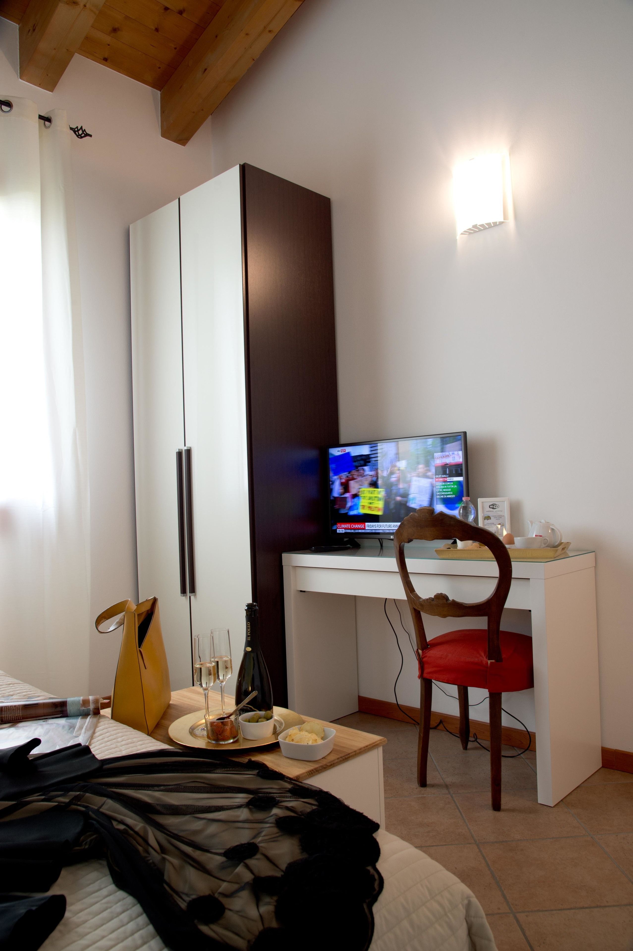 A hotel room with a bed, a desk with a computer, a chair, a wardrobe, and a small table with a bottle and glasses.