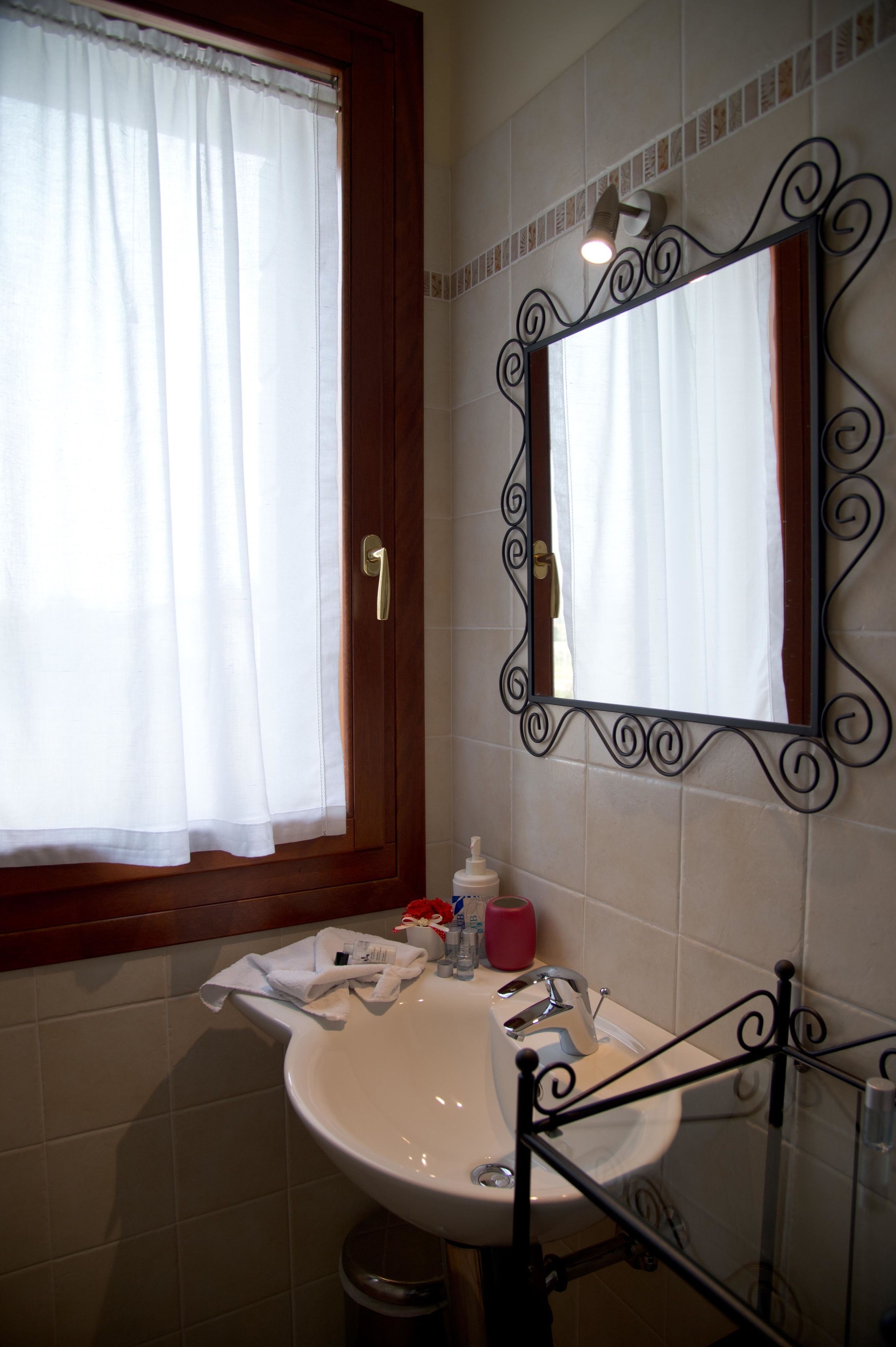 Ein Badezimmer mit weißem Waschbecken, einem Spiegel mit Zierrahmen, einem Fenster mit Vorhang und einem Handtuchhalter.