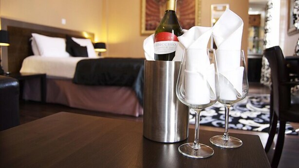 A hotel room with a bed, a bedside table with a wine bottle and glasses, and a nightstand with a lamp. - Villaggio Hotel & Restaurant (Warrington)