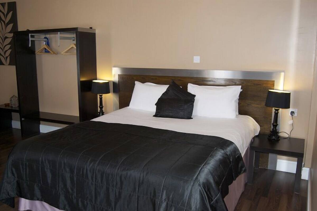 A neatly made bed with a black bedspread, two white pillows, and a wooden headboard. There are two bedside tables with lamps, and a coat rack with hangers to the left.