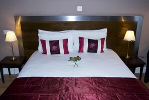 A neatly made bed with white and maroon bedding, two bedside lamps, and a single rose on the bed. - Villaggio Hotel & Restaurant (Warrington)