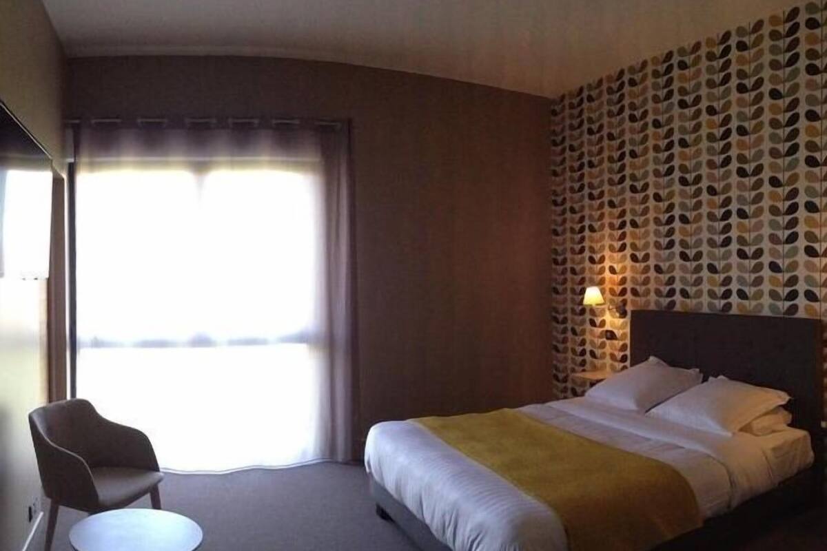 A hotel room with a bed, a chair, a small table, and a patterned wall.