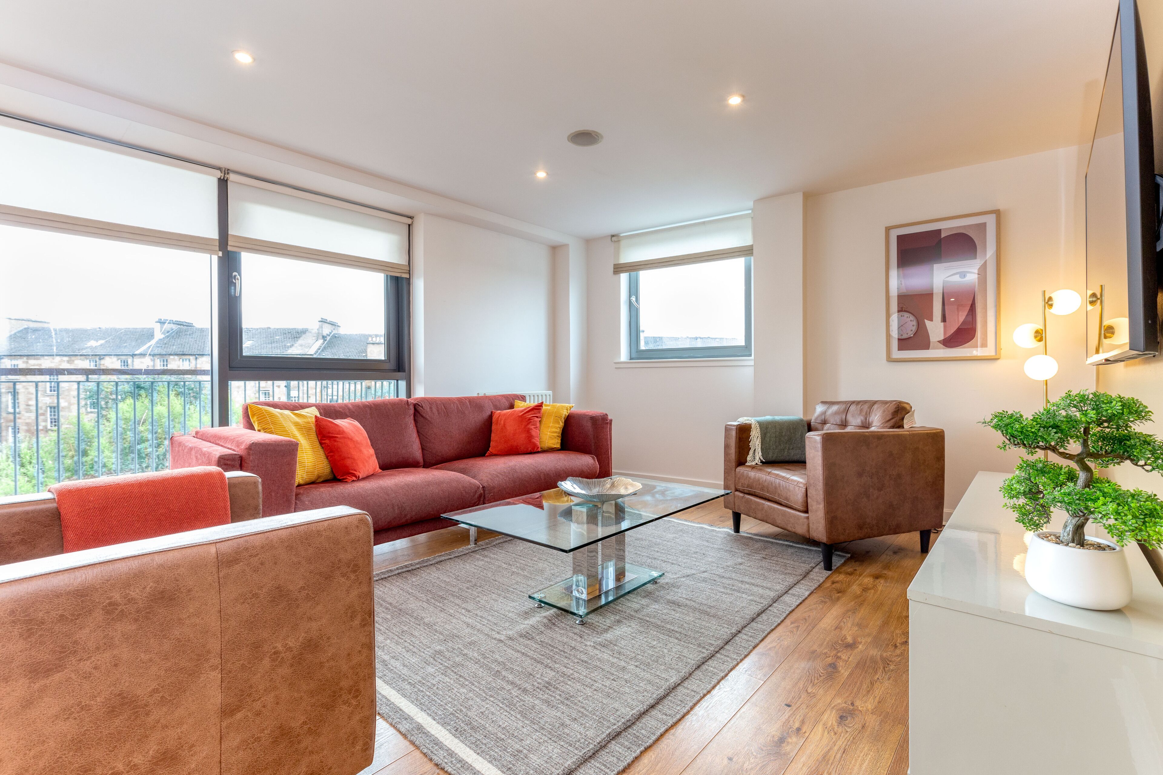 A modern living room with a glass coffee table, red and brown sofas, a potted bonsai tree, and large windows offering a city view.