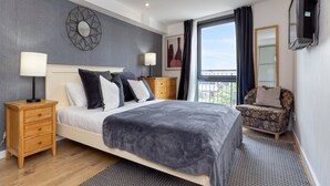 A modern bedroom with a large bed, a wooden nightstand, a patterned armchair, and a view of the city through a large window.