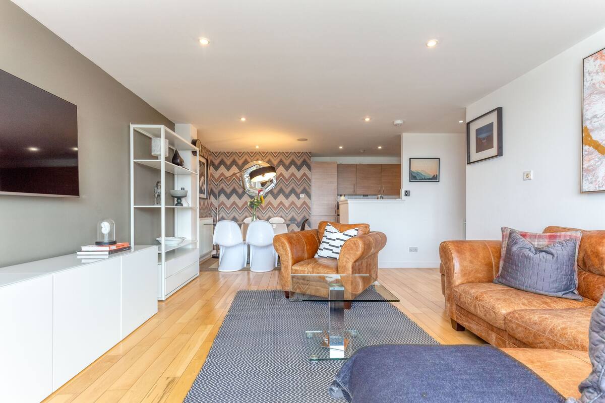 A modern living room with a sofa, a glass coffee table, and a TV mounted on the wall. There is a kitchen area in the background with cabinets and a countertop.