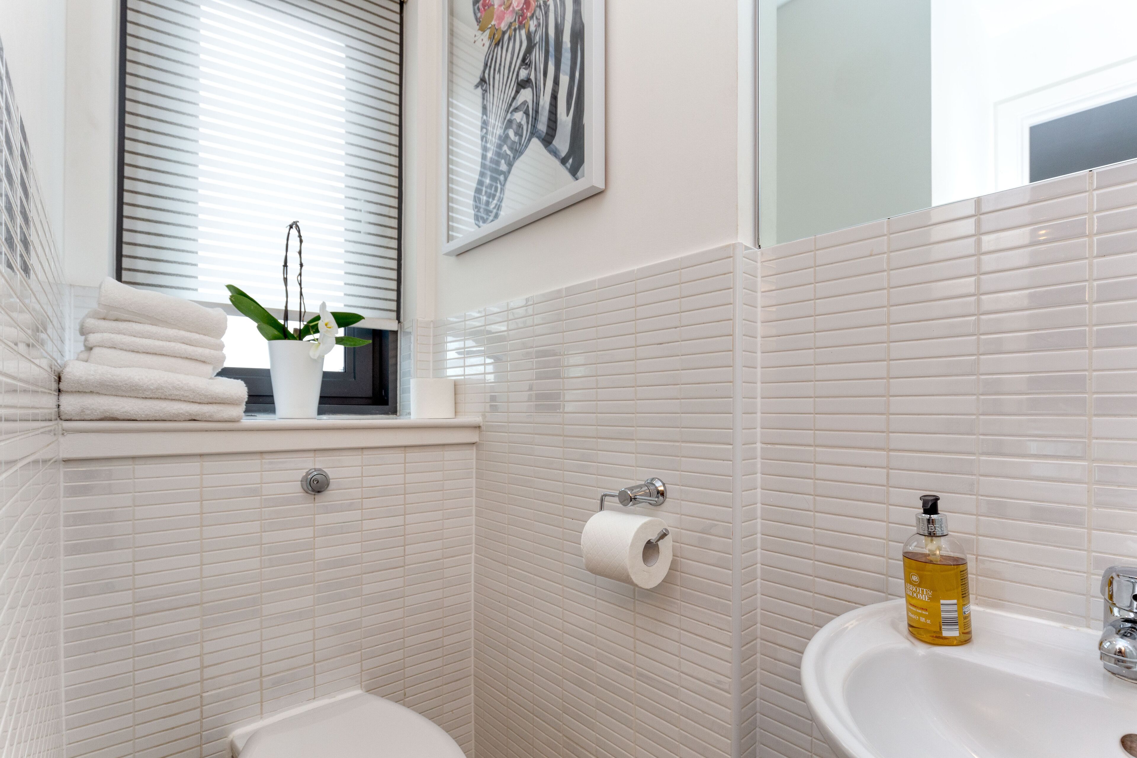 Un baño con un lavamanos blanco, un inodoro y una ventana con persianas. Hay un perchero con toallas y una planta en el alféizar.