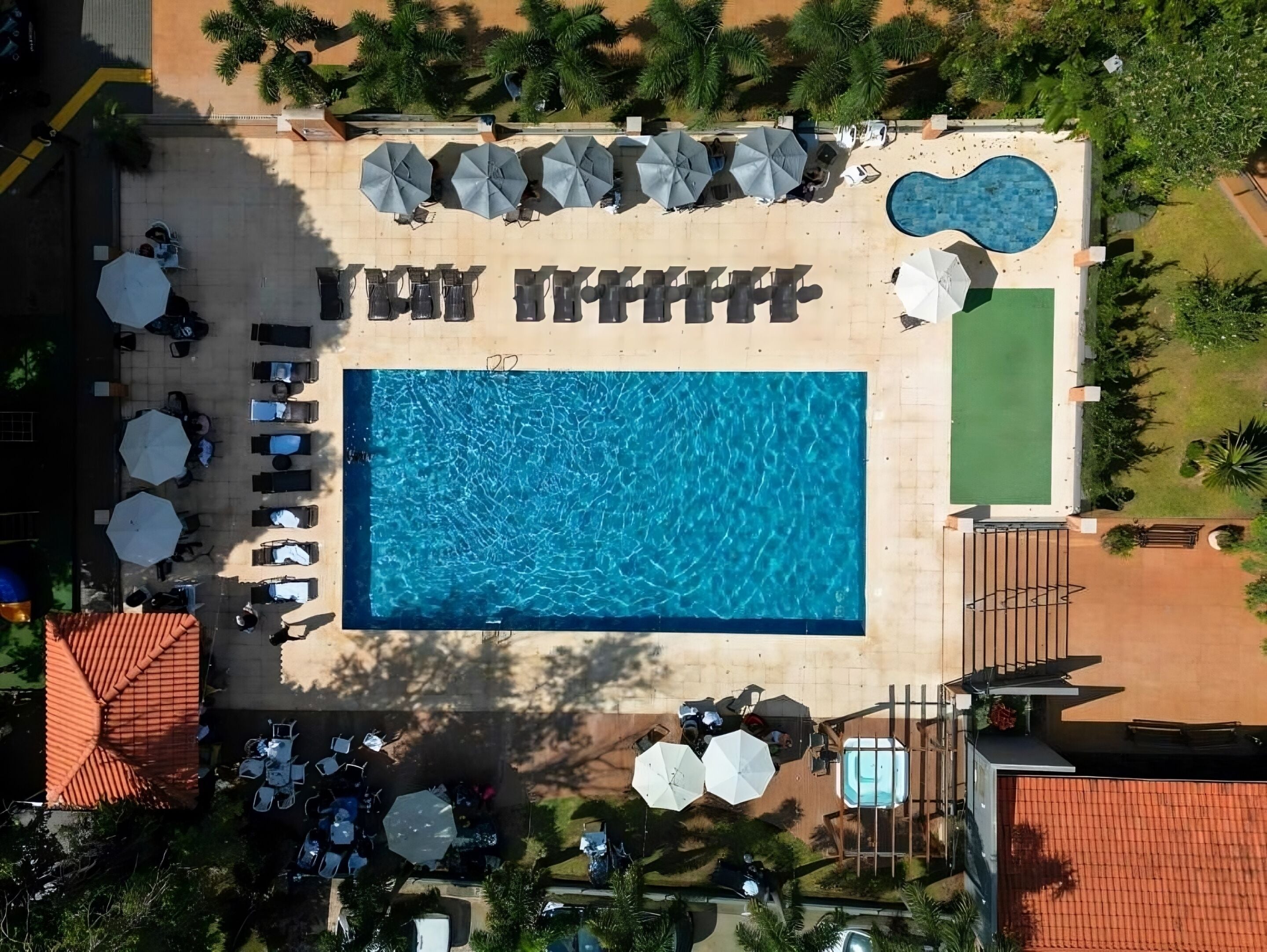An aerial view of a resort with a rectangular swimming pool surrounded by lounge chairs and umbrellas, a separate smaller pool, and a fenced area with a hot tub.
