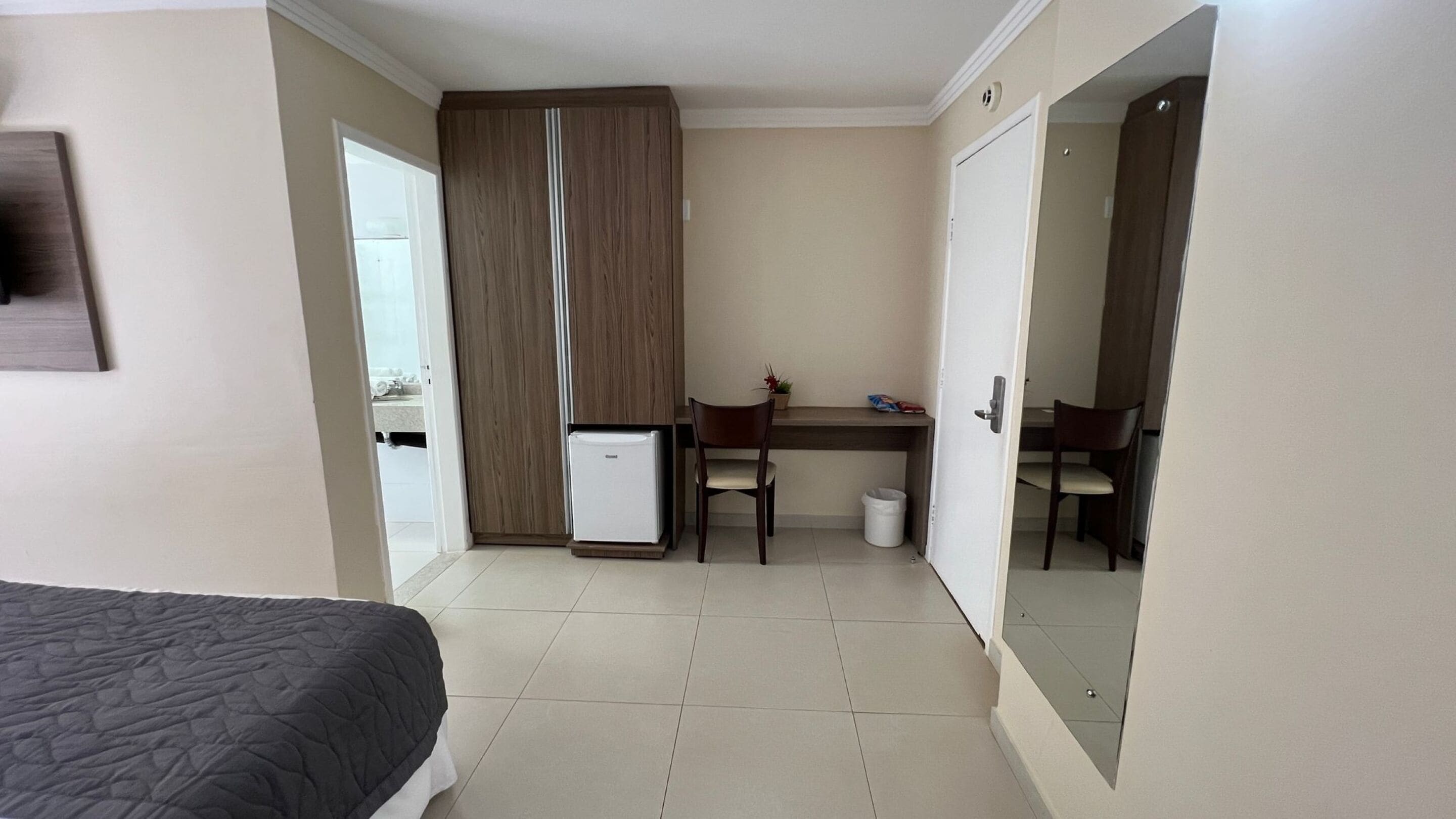 Quarto de hotel com cama, escrivaninha, cadeira, um pequeno frigorífico e espelho.