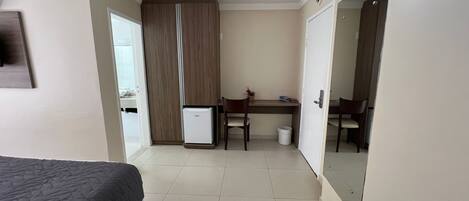 Quarto de hotel com cama, escrivaninha, cadeira, um pequeno frigorífico e espelho.