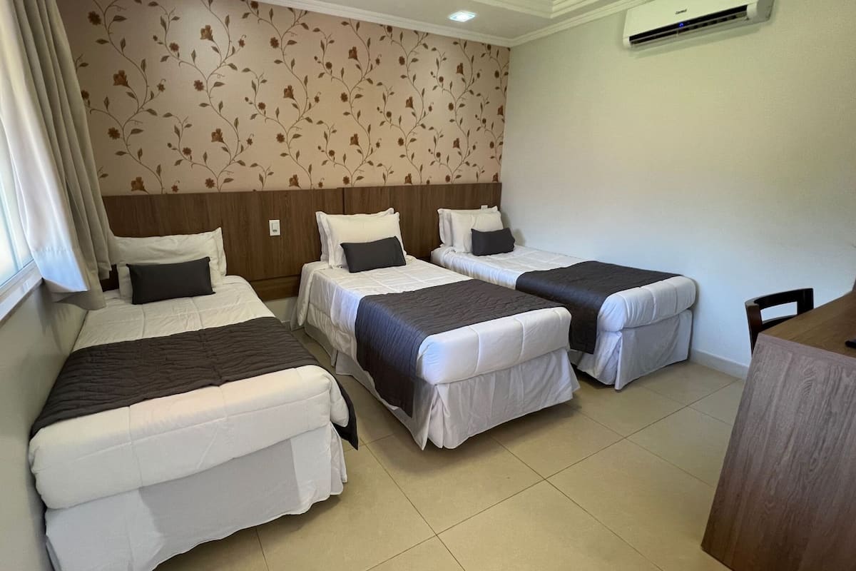 A hotel room with three single beds, a wooden headboard, a window with curtains, and an air conditioning unit on the wall.