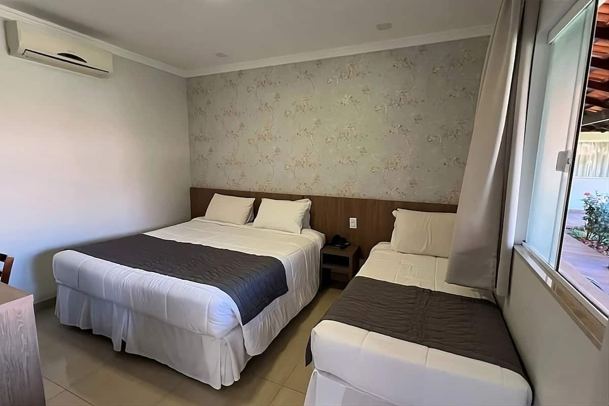 A hotel room with a large bed, a smaller bed, a nightstand, an air conditioning unit, and a window with a view of the outside.