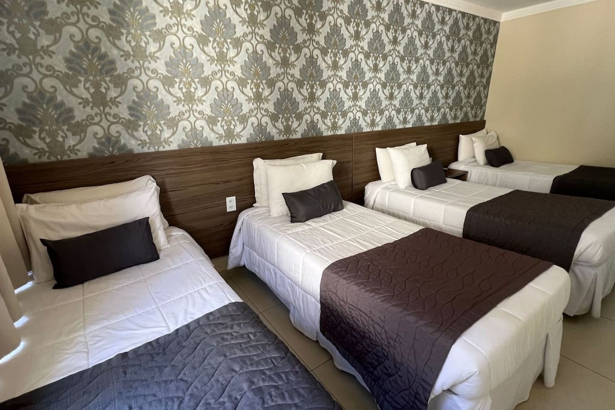 A hotel room with four single beds, each with a white duvet and a dark pillow. The room has a wooden headboard and a patterned wallpaper.