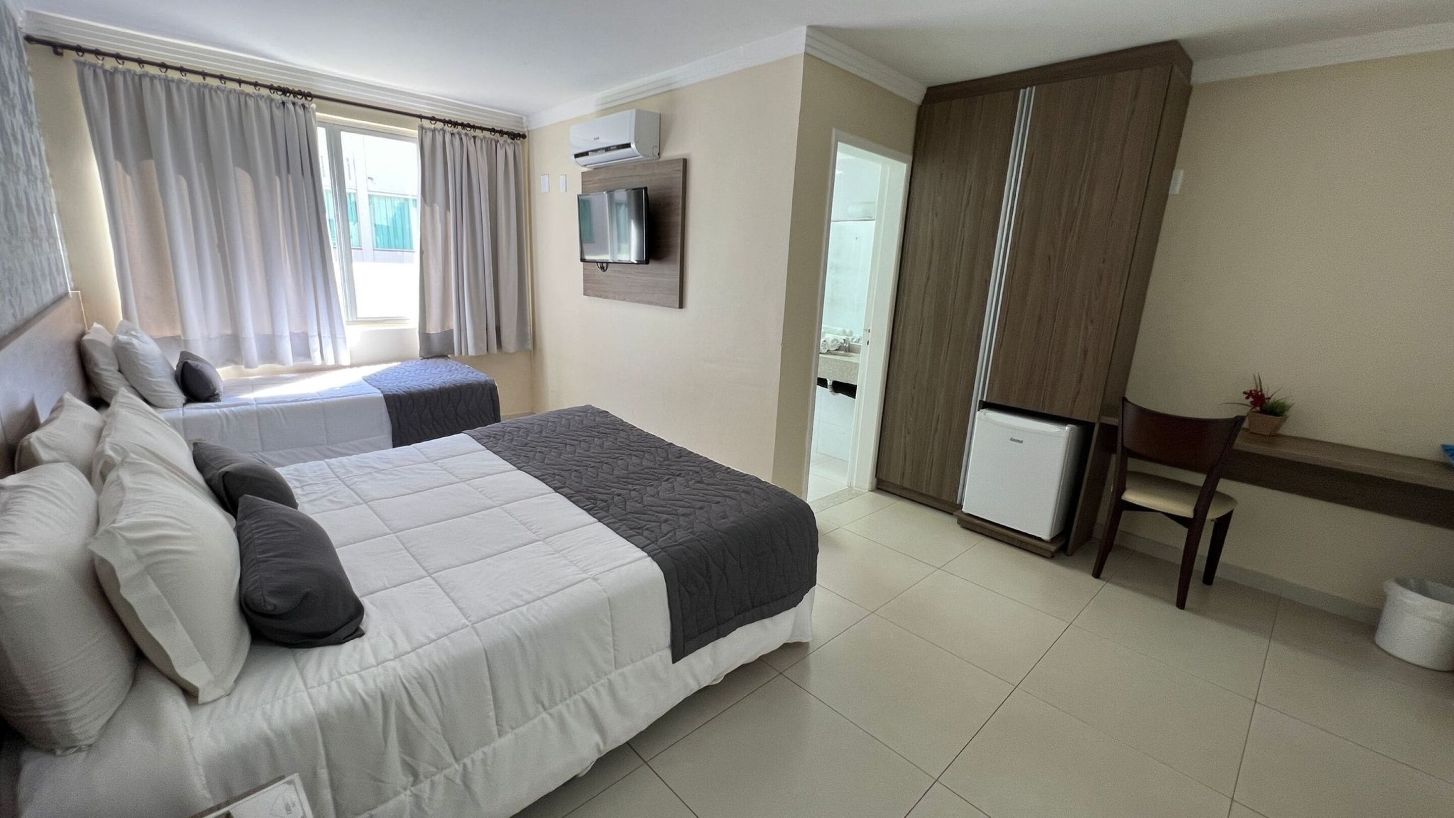 Quarto de hotel com duas camas, televisão, mini-geladeira, escrivaninha e janela com cortinas.