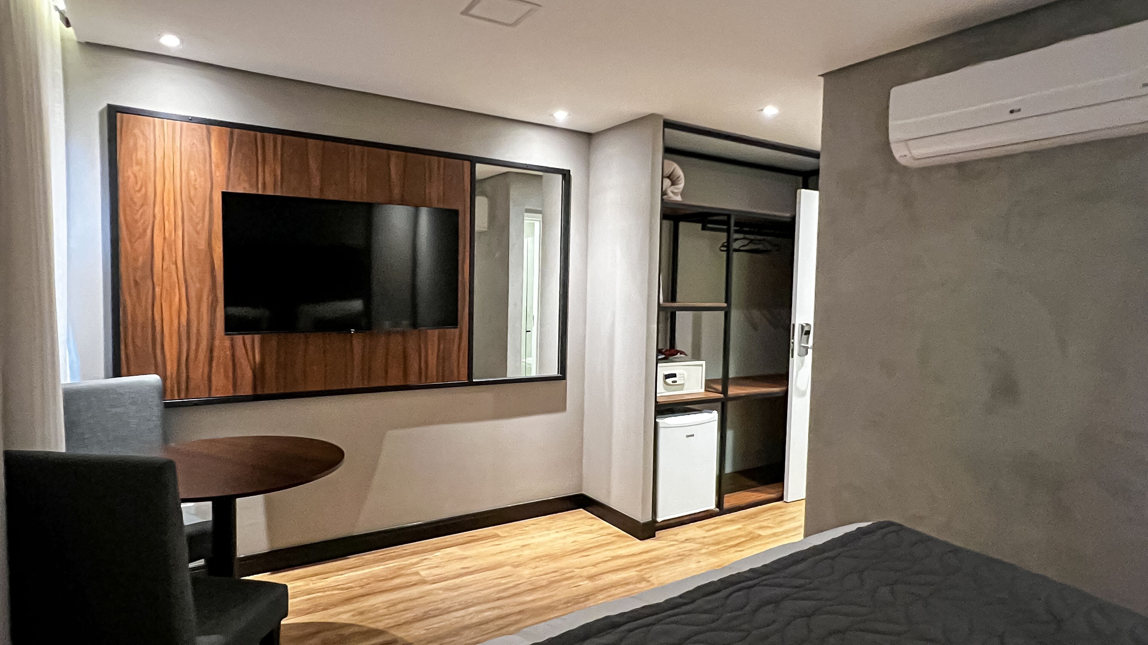 A modern hotel room with a wooden floor, a flat-screen TV mounted on a wooden panel, a small round table, a bed, and a wardrobe with a mirror.