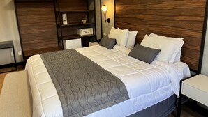 A modern hotel room with a large bed, a bedside table, a nightstand, a built-in shelf, and a wall-mounted lamp.