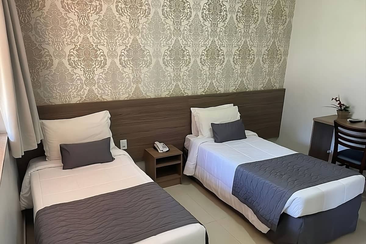 A hotel room with two beds, a wooden headboard, a nightstand with a phone, and a small desk with a plant.