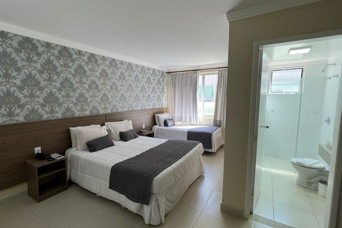 A hotel room with two beds, a wooden headboard, a nightstand, a bathroom with a toilet and shower, and a patterned wallpaper.