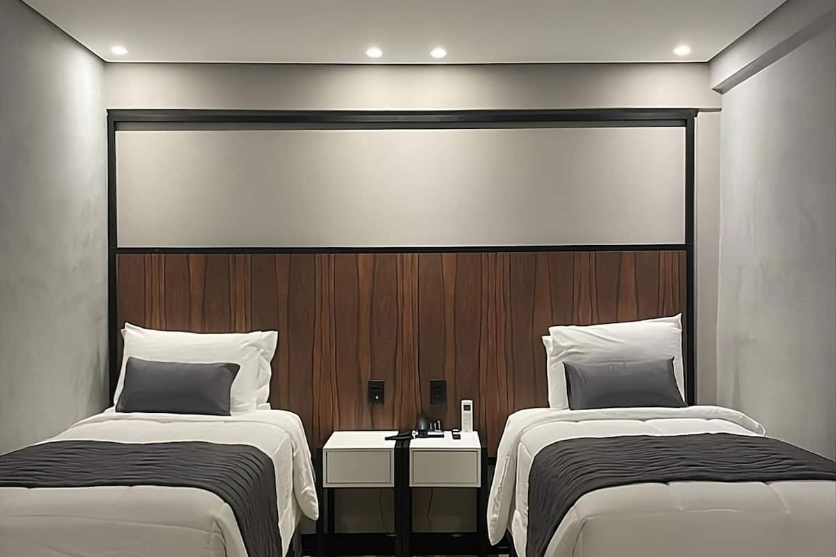 A modern hotel room with two beds, a wooden headboard, a small bedside table with a lamp, and a wooden floor.