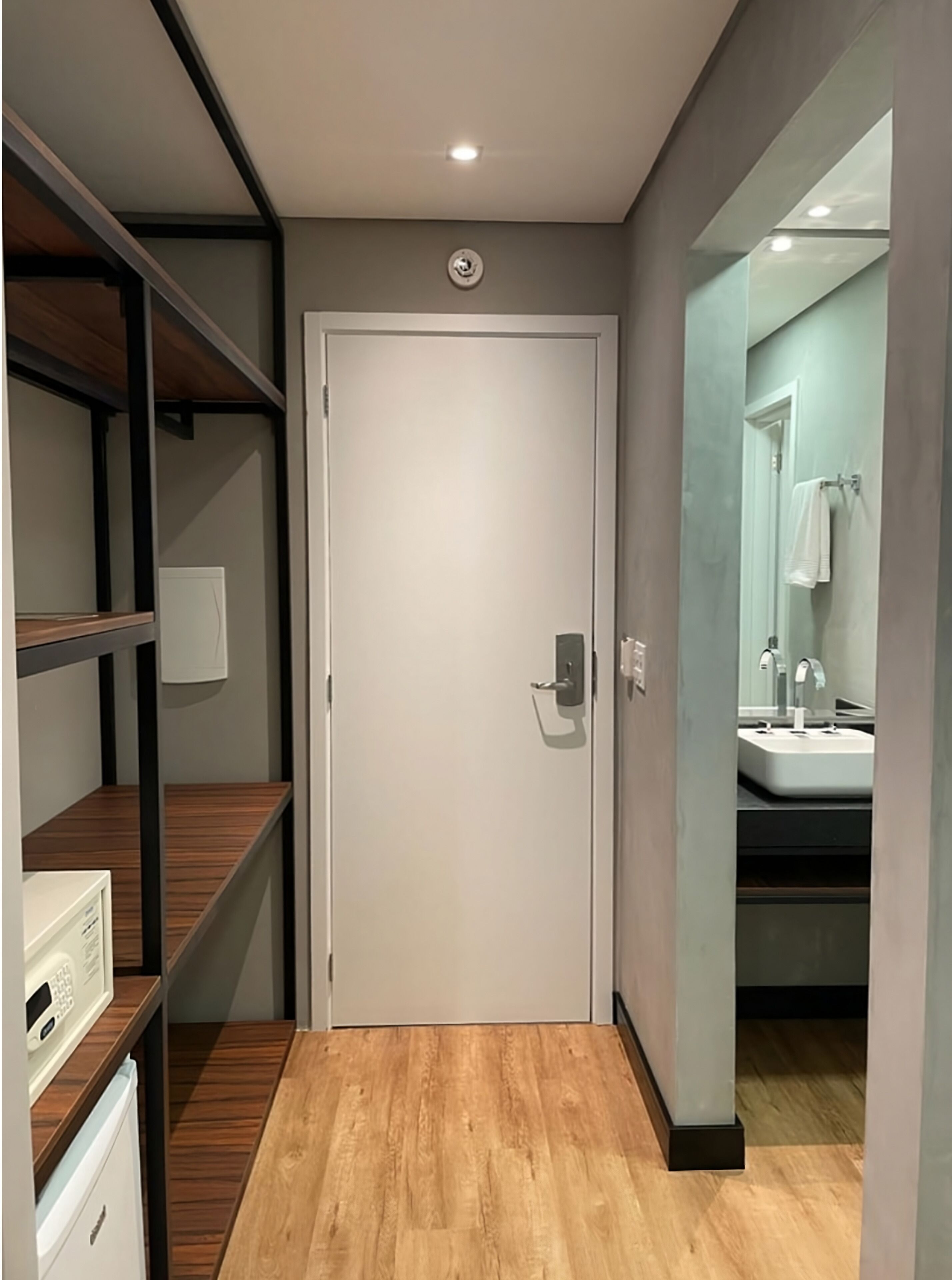 A narrow hallway with a white door, wooden shelves, a microwave, and a sink visible through a glass partition.