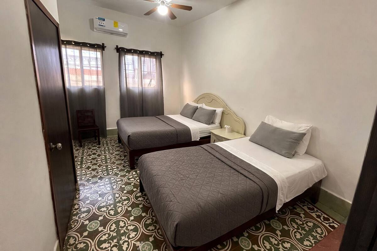 A hotel room with two beds, a ceiling fan, an air conditioning unit, and patterned floor tiles.