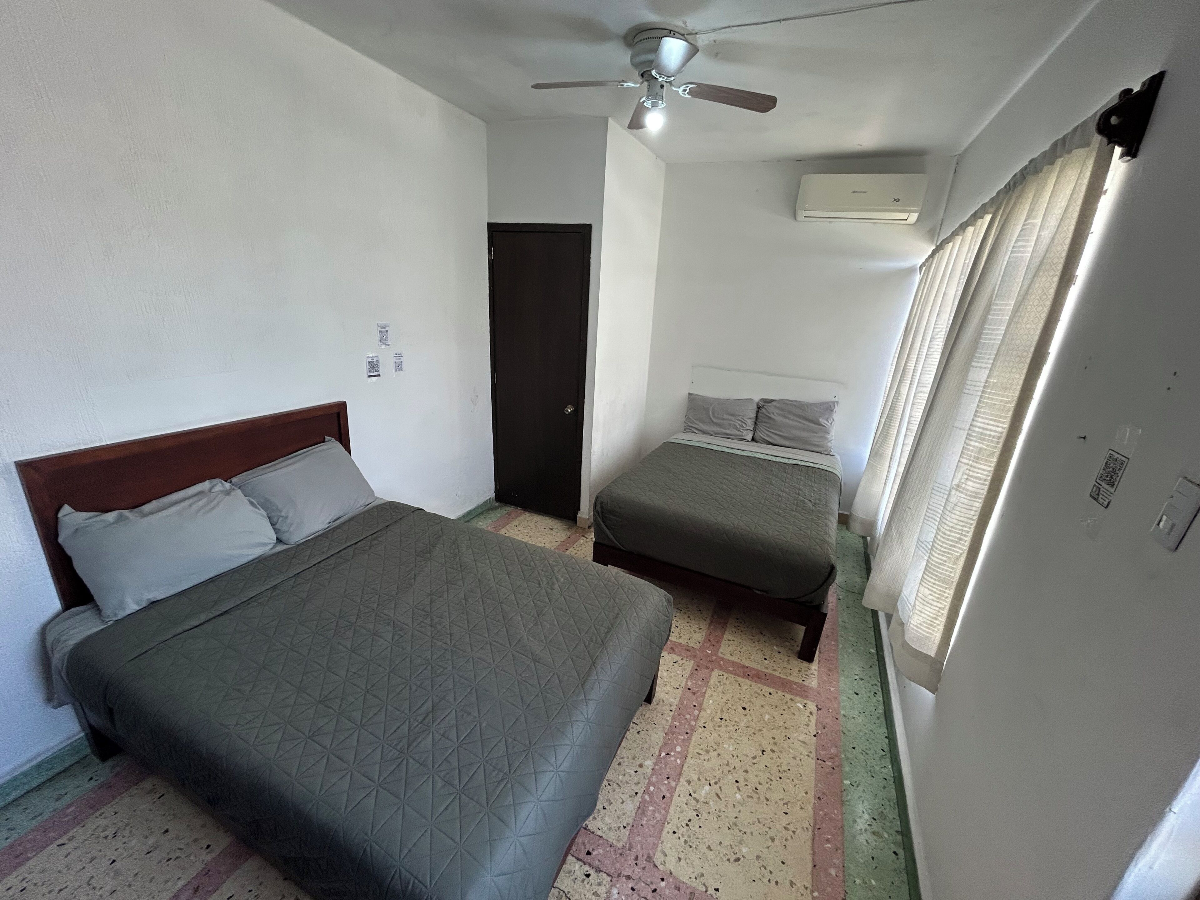Une petite pièce propre, avec deux lits, un ventilateur de plafond, un appareil de climatisation et une porte.