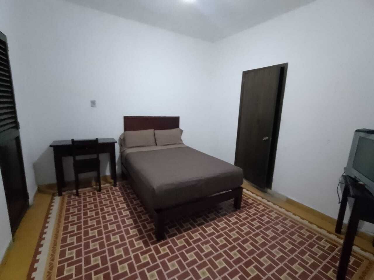 Una habitación pequeña con una cama individual, cabecera de madera, una mesita y un televisor sobre un soporte.