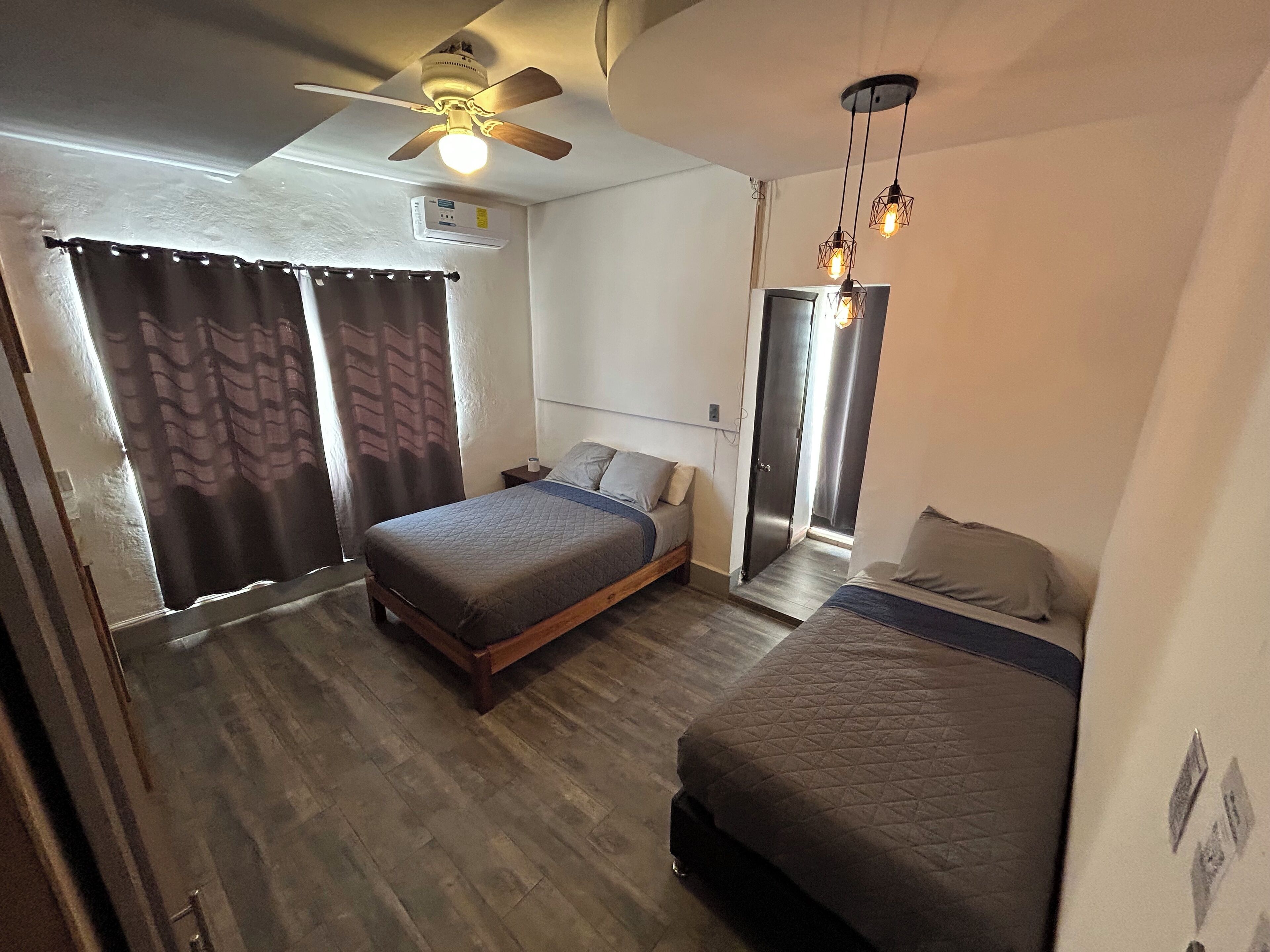 Een slaapkamer met twee bedden, een plafondventilator, een airconditioning en een deur naar een andere kamer.