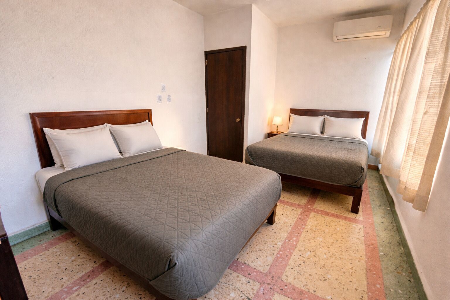 Habitación de hotel con dos camas, una puerta, un equipo de aire acondicionado y una ventana con cortinas.