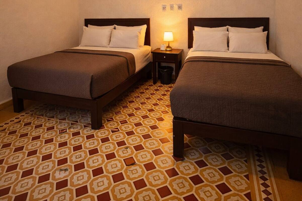 A hotel room with two beds, a ceiling fan, a patterned floor, and a nightstand with a lamp.