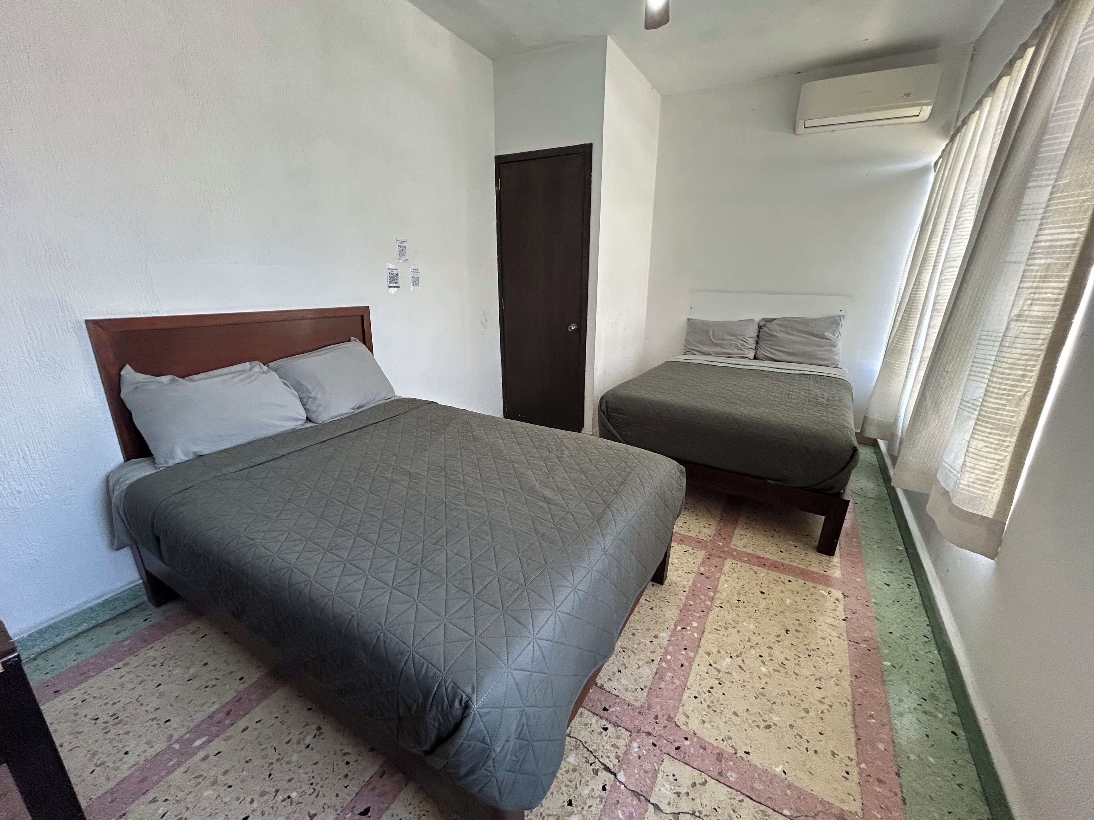 A bedroom with two beds, a door, an air conditioning unit, and a window with curtains.