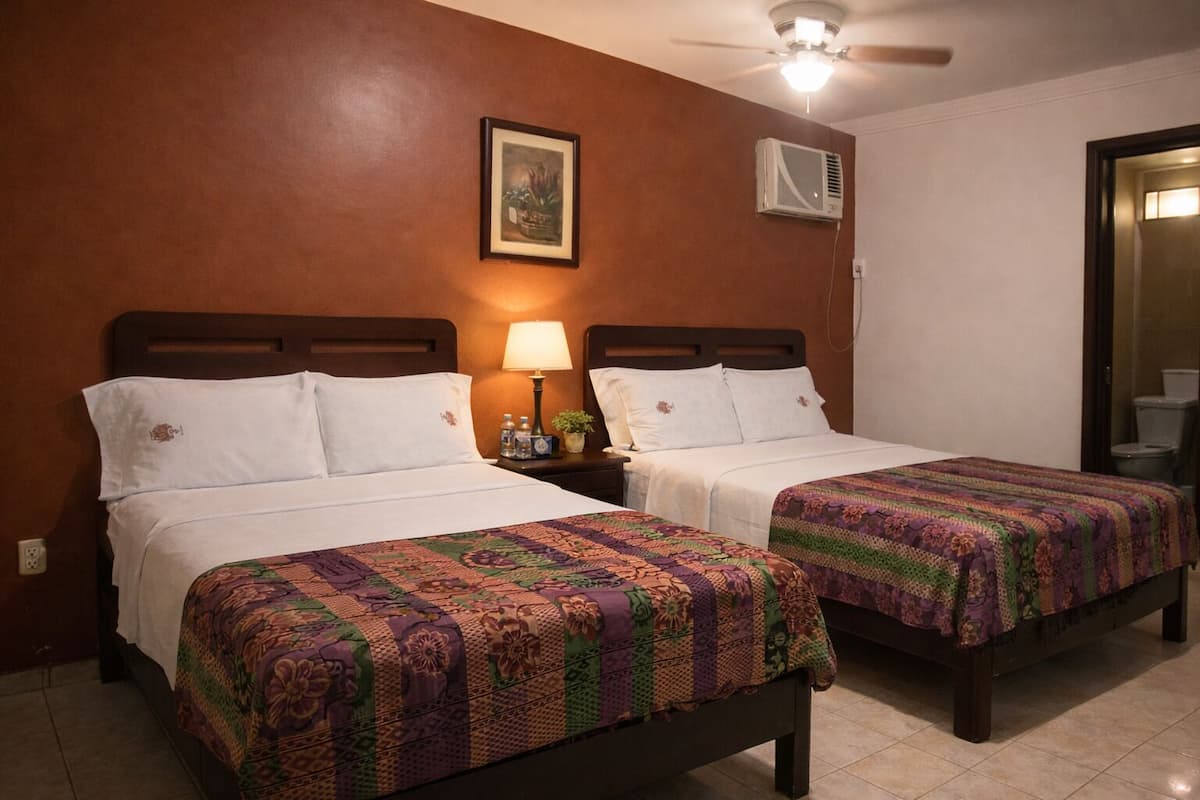 Habitació d'hotel amb dos llits, un ventilador de sostre, un aparell d'aire condicionat, un quadre encimbellat a la paret i una taula de nit amb una làmpada i ampolles d'aigua.