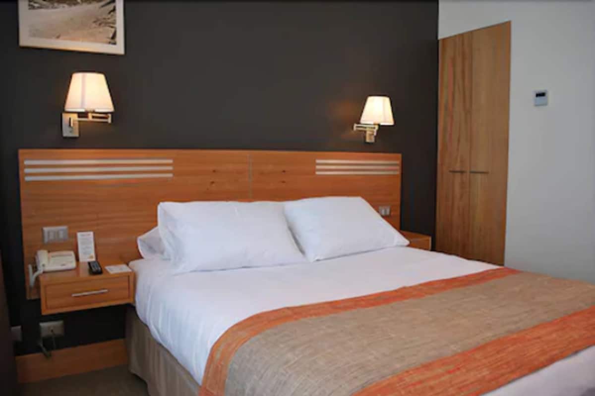 A hotel room with a wooden headboard, two bedside lamps, a telephone, and a bed with a beige and orange blanket.