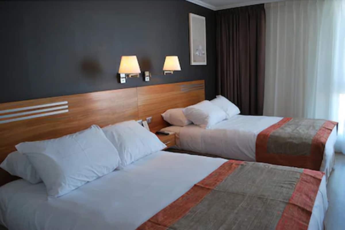 A hotel room with two beds, a wooden headboard, wall-mounted lamps, and a large window with curtains.