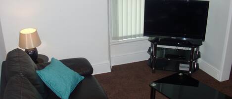 A living room with a black leather sofa, a glass coffee table, a flat-screen TV on a stand, a lamp, and a window with blinds.