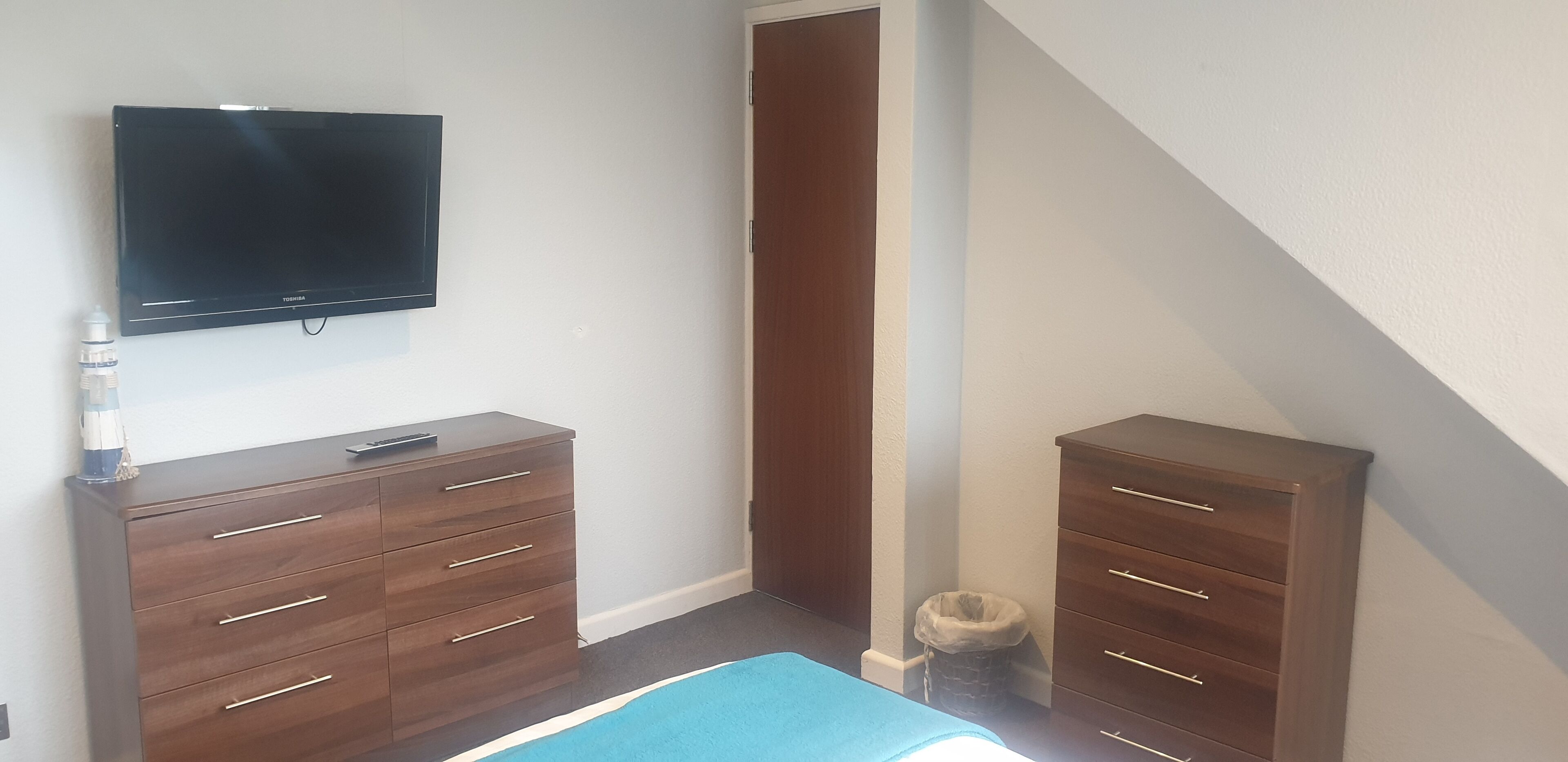 A bedroom with a TV mounted on the wall, two wooden dressers, a blue rug, and a small trash can.