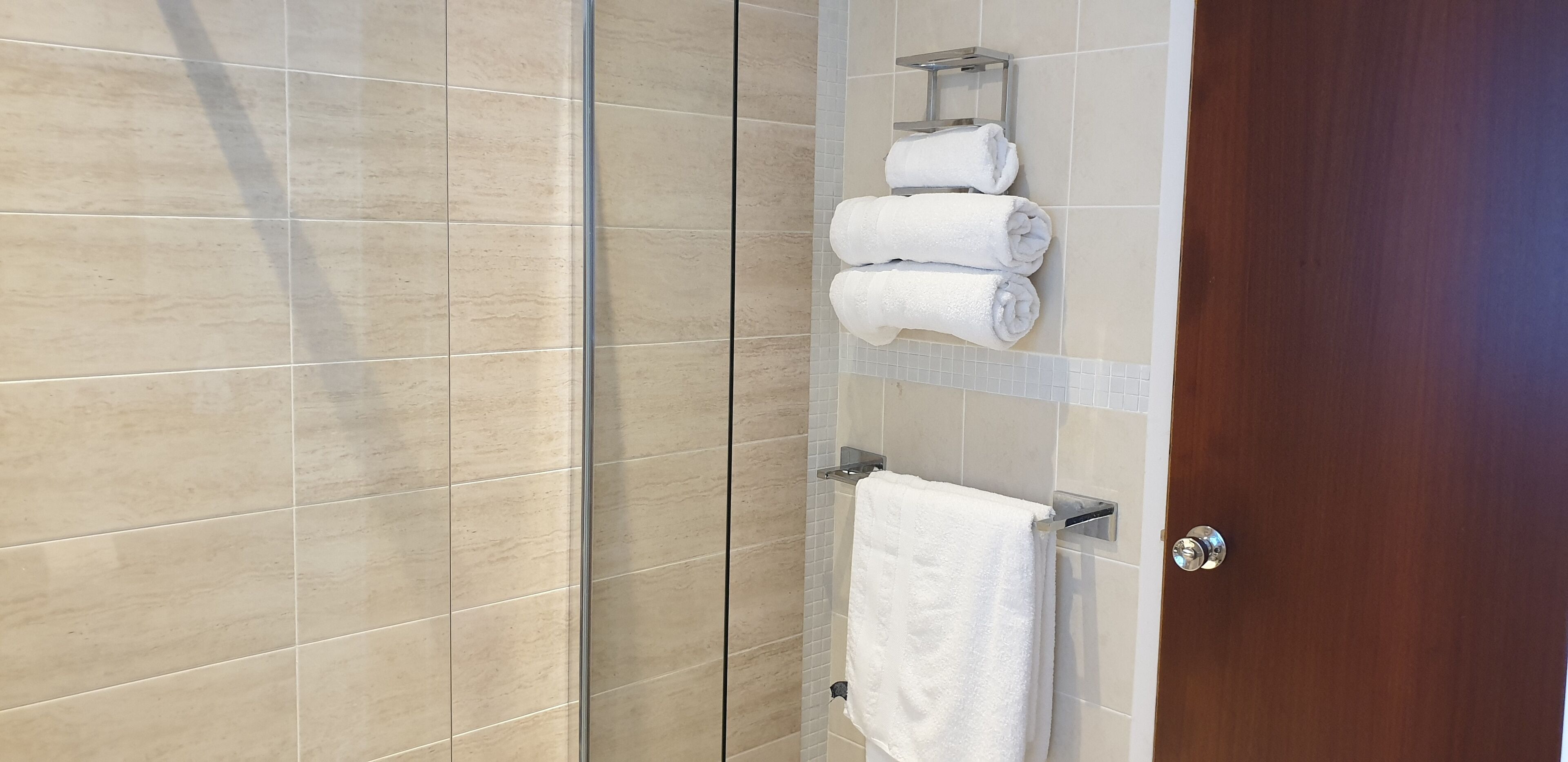 A bathroom with beige tiles, a wooden door, and a towel rack holding three rolled towels.