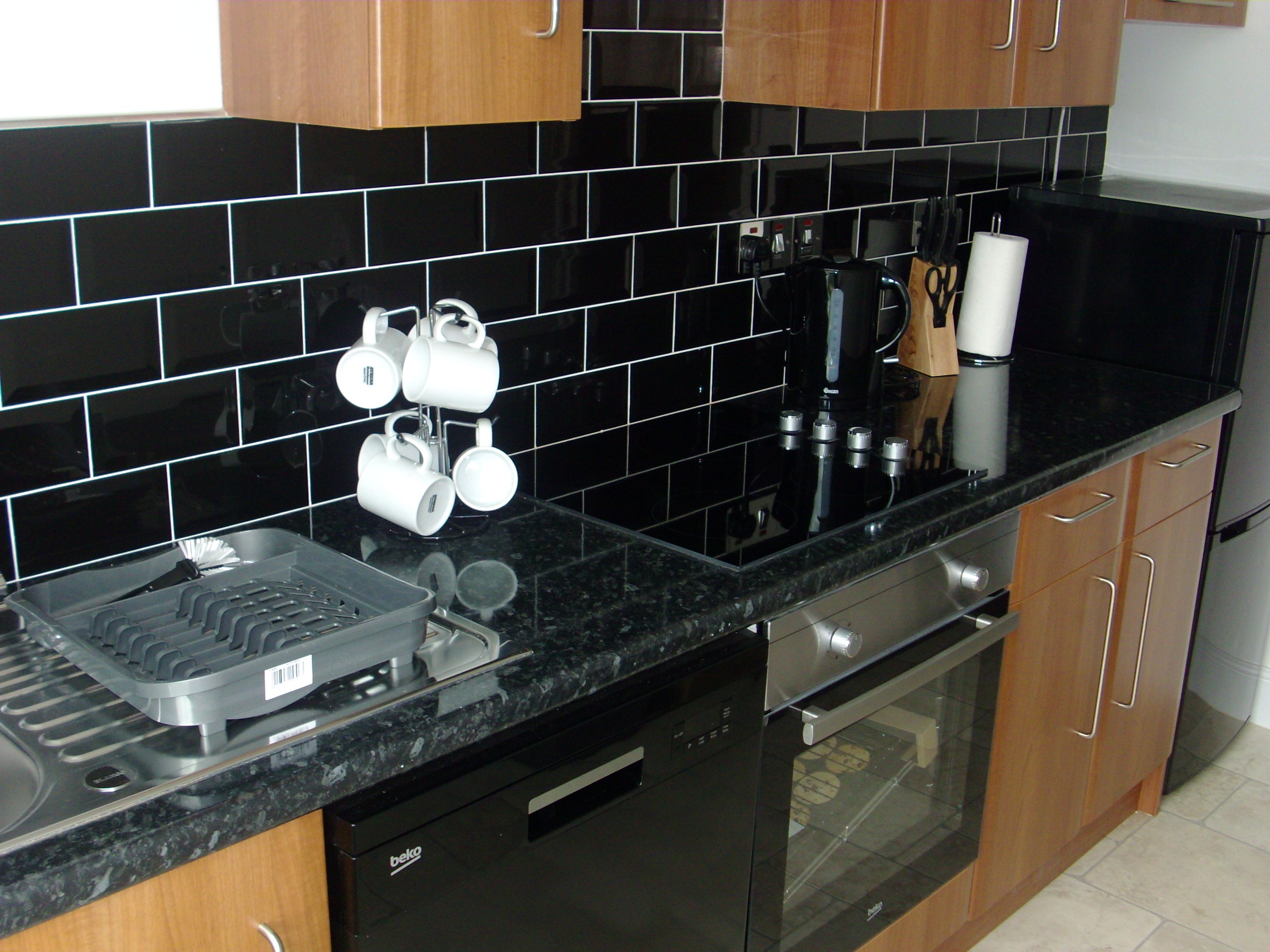 A kitchen with black tiled backsplash, wooden cabinets, a black countertop, a coffee maker, and a dish rack.